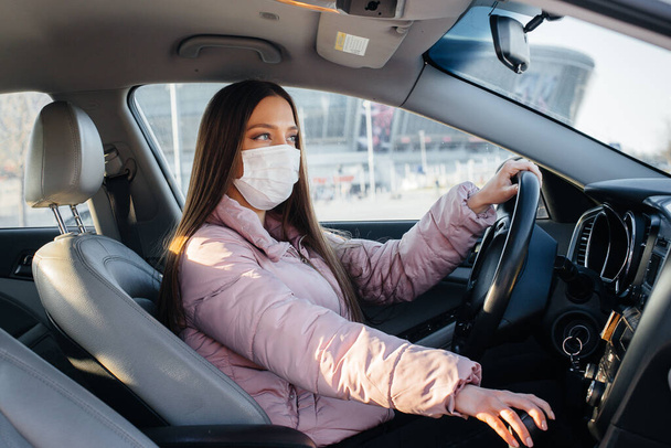 A young girl sits behind the wheel in the car in the mask during the global pandemic and coronavirus. Quarantine - 写真・画像