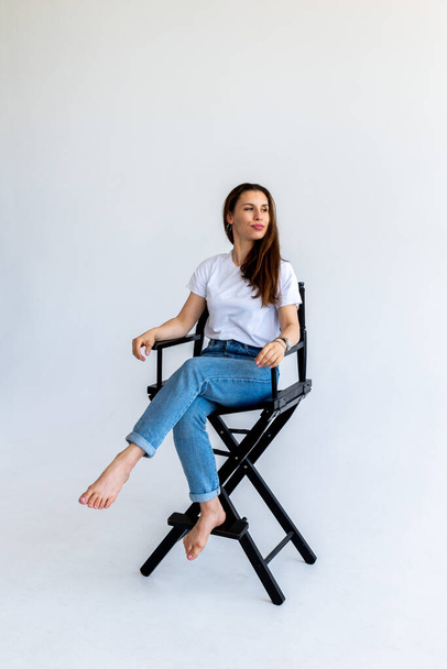 Adult woman sits on a filmmaker's chair in blue jeans and a white T-shirt. Red hair in humans. White background - Fotografie, Obrázek