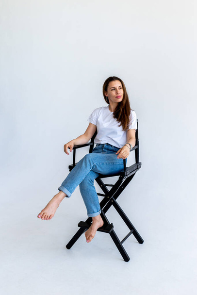 Adult woman sits on a filmmaker's chair in blue jeans and a white T-shirt. Red hair in humans. White background - Fotoğraf, Görsel