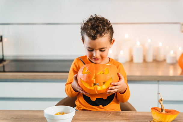 Child dressed in an orange pumpkin shirt holding and looking at his pumpkin decorated and lit with a candle to celebrate halloween night - Fotó, kép