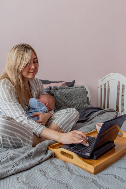 Young woman in comfortable home clothes works on laptop with a baby in her arms in bedroom. Home wellbeing concept. Emotional health mom woman - Zdjęcie, obraz