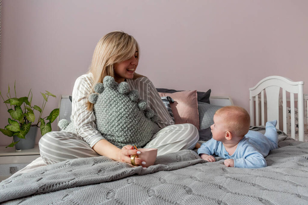 Young woman sits on bed in bedroom and drinks coffee next to her is infant. Good morning. Wellness concept - Fotografie, Obrázek