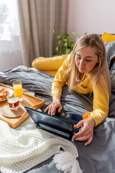 Young woman in yellow pajamas has healthy breakfast and work laptop while lying on bed in bedroom. Home wellbeing concept. Emotional health of a young woman. Wellness - Фото, зображення