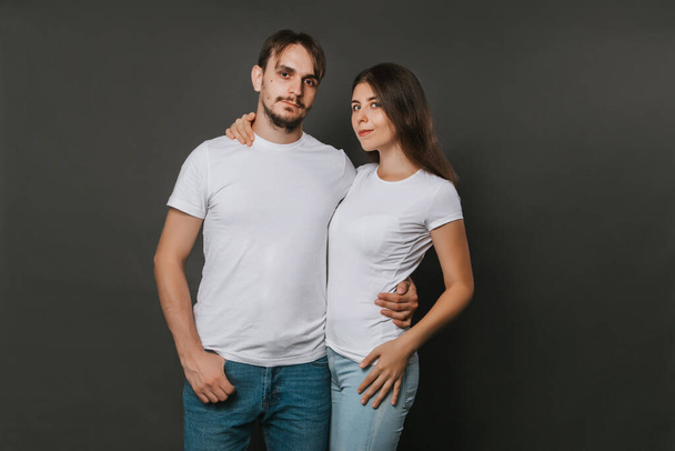 Ein junges Paar, eine Frau und ein Mann in weißen Blanko-T-Shirts vor dunkelgrauem Hintergrund. Attrappe. - Foto, Bild