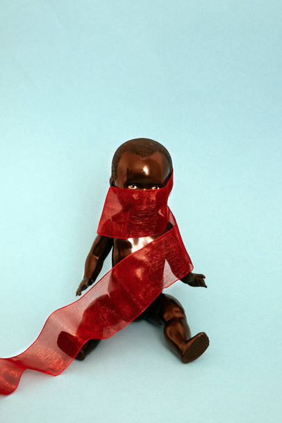 black doll covered with a red ribbon sitting on blue background - Foto, Imagem