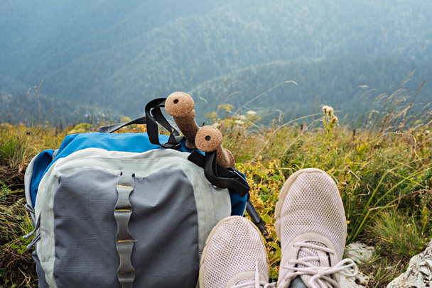 Hiking backpack, trekking poles and legs in sports shoes of young woman resting in a mountain hike in the valley, active healthy lifestyle outdoor recreation - Photo, Image