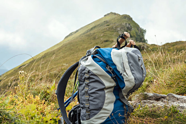 大きな灰色の青いハイキングバックパック、山のピークの背景にある緑の植物の間のトレッキングポール、ハイキングアクティブな健康的なライフスタイル屋外レクリエーション - 写真・画像