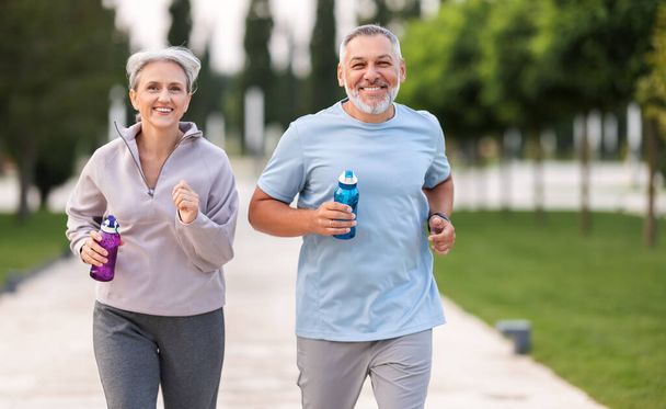 Jolies retraités joyeux couple jogging à l'extérieur dans le parc de la ville le long de ruelle avec des arbres verts, mari heureux et femme qui se regardent avec le sourire tenant des bouteilles d'eau dans les mains - Photo, image