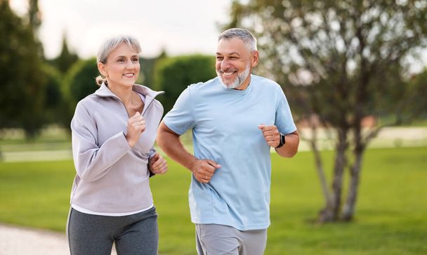 Happy senior husband and wife in sportive outfits running outdoors in city park, lovely retirees couple jogging in sunny morning looking at each other with warmth and smile. Healthy lifestyle concept - Photo, Image