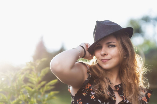 Attractive young girl wearing a black hat is posing outdoors looking at the camera. - Φωτογραφία, εικόνα