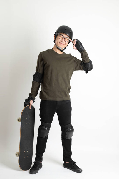 Азіатський чоловік з захисним спорядженням і скейтбордом стоїть на білому фоні.. - Фото, зображення