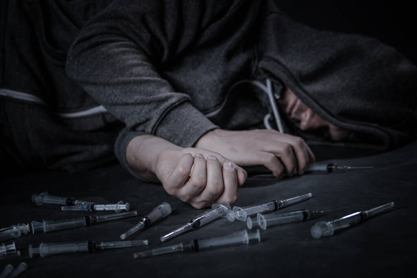 A drug addicted person draws a dose of the drug in a syringe. The guy is an addict, the concept of the fight against drugs. A young man sits in a pile of used syringes. - Photo, image