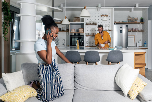 Young mixed-race couple in their home a black African woman talks on the phone and is happy because she got good news and a man stands in the kitchen and laughs while cooking lunch - Foto, Bild