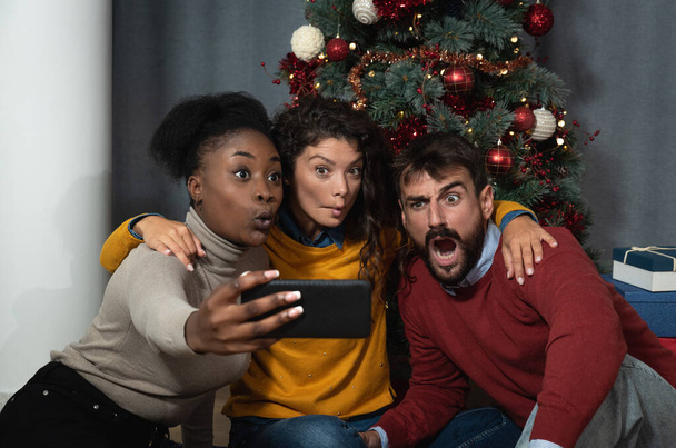 Three young friends celebrate Christmas and New Year's Eve together by joking and taking selfies with a smartphone making funny facial expressions in front of the Christmas tree - Foto, Imagem