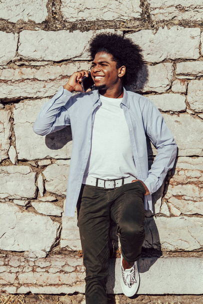 junger schwarzer Mann mit Afro spricht auf Handy an Wand gelehnt - Foto, Bild