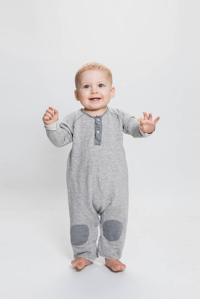 Cute little baby boy in grey clothes standing on the White Background. The child learns to stand and walk independently.  Studio light. Isolated. Caucasian kid 1 year old with blue eyes. - 写真・画像