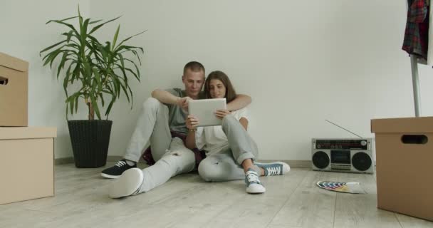 Jong stel in nieuwe flat met behulp van tablet. Man en vrouw in de twintig zitten op de vloer van een nieuw appartement met digitale tablet. Real-time, hand-held schot, medium schot, 4K. - Video