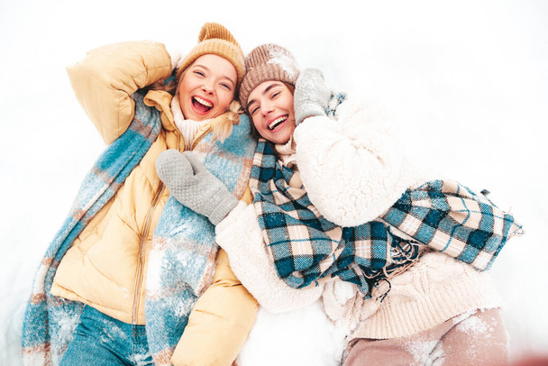 Two young beautiful smiling hipster female in trendy warm clothes and scarfs.Carefree women posing in the street in park. Positive pure models lying in snow. Enjoying winter moments. Xmas concept - Фото, изображение