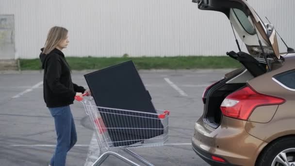Nainen kuljettaa modernia suurta televisiota supermarketin vaunussa ja yrittää ladata sen auton takakonttiin pysäköintipaikalla. - Materiaali, video