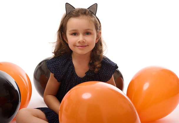 Portrait of cute witch girl in Halloween dress and hoop with cat ears having fun with colorful black and orange balloons isolated on white background with copy space. Halloween party. - Fotó, kép