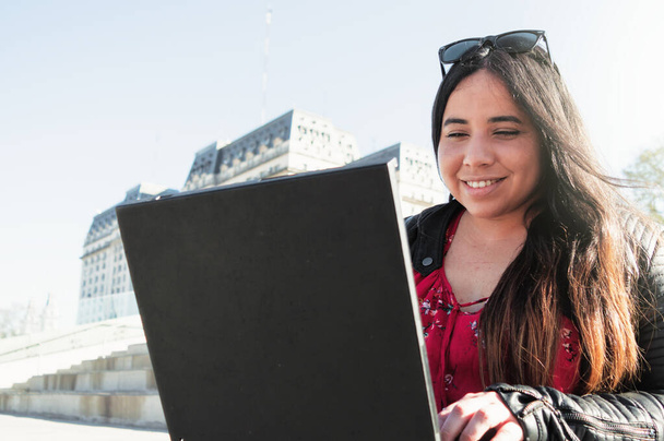 mooie jonge witte latino freelancer zakenmeisje met behulp van haar computer dragen van een zonnebril, rood en zwart kleren glimlachen gelukkig buiten, hemel op de achtergrond - Foto, afbeelding