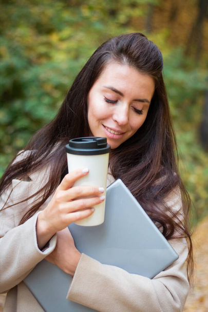 Brunette woman in autumn park with laptop and cup of coffee. Student. Teacher. Online education. - Zdjęcie, obraz