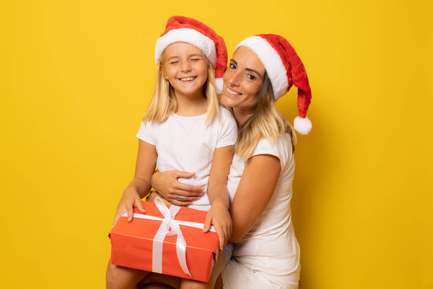 黄色の背景の上にサンタの帽子の幸せな母親と娘。クリスマスシーズンのコンセプト. - 写真・画像
