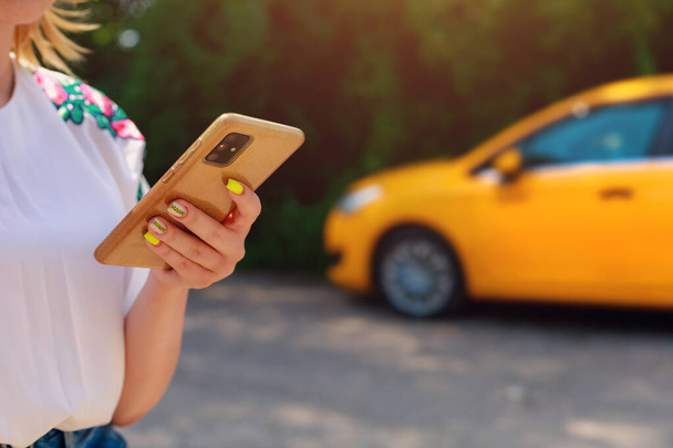 Smartfon lub telefon w kobiecych rękach na rozmytym tle żółtego taksówki. Połączenie samochodowe i kontrola we wniosku. - Zdjęcie, obraz