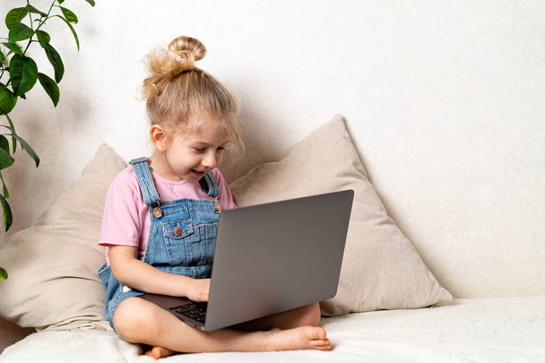 Joyeux petite fille blonde est assise à la maison sur un lit avec un ordinateur portable, un endroit pour le texte, le concept d'apprentissage à la maison. - Photo, image