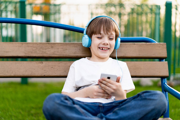 a happy little boy on the street sits on a bench with headphones and a phone and smiles.  - Photo, Image