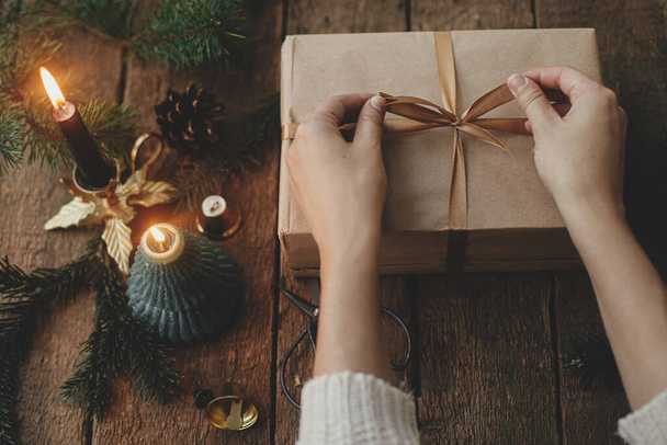 Ręce owijania stylowy prezent na Boże Narodzenie w papier rzemieślniczy na rustykalnym drewnianym tle ze świecą, nożyczki, gałązki jodły. Nowoczesne proste przyjazne dla środowiska xmas teraźniejszość, skandynawski nastrój obraz - Zdjęcie, obraz