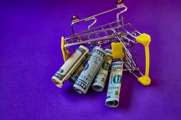 concept - curled dollar banknotes fall out of Shopping Cart Trolley against the purple background - Фото, изображение