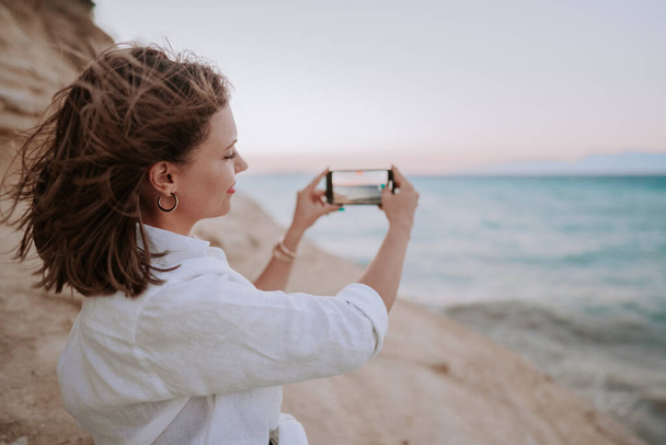 Young woman filming video with mobile phone on beach near blue sea. Girl using smartphone, taking photos for social app, enjoying vacation time. - Zdjęcie, obraz