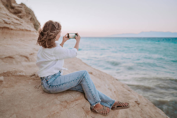 Young woman filming video with mobile phone on beach near blue sea. Girl using smartphone, taking photos for social app, enjoying vacation time. - Fotografie, Obrázek
