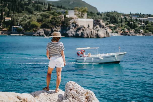 Woman in striped cruise outfit is watching on motor boat is sailing alongside. Lady in straw hat standing on berth. Blue sea background. Riviera coast, luxury vacation concept. - Fotó, kép