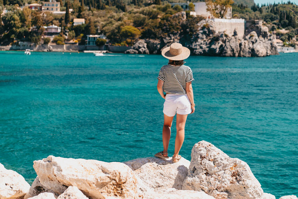 Back view of woman in striped cruise outfit, straw hat. Lady standing on stone beach. Teal sea background. South Europe Riviera, travel destination, luxury vacation concept. - Photo, Image
