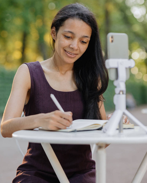 young millennial african american woman watching a webinar or live stream from her smartphone and taking notes in her notebook - Fotografie, Obrázek