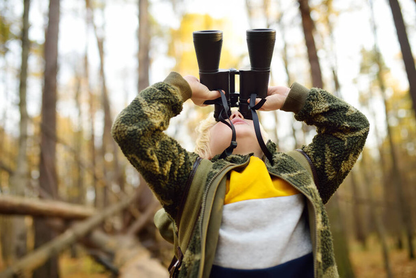 Little boy scout with binoculars during hiking in autumn forest. Child is sitting on large fallen tree and looking through a binoculars. Concepts of adventure, scouting and hiking tourism for kids. - Φωτογραφία, εικόνα