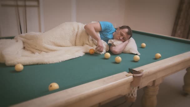 De man verveelt zich, ligt op de pooltafel en rolt ballen op tafel, bedekt met een deken - Video