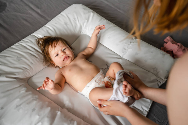 Small caucasian baby girl or boy lying on the bed in bright room while hands of her mother changing diapers and clothes copy space parenting childhood concept getting dressed top view - Photo, Image