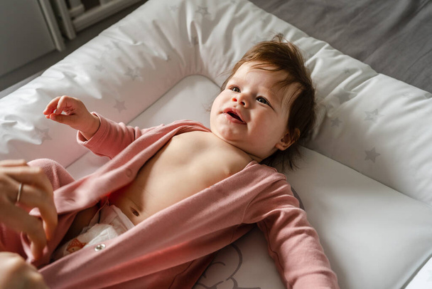 Small caucasian baby girl or boy lying on the bed in bright room while hands of her mother changing diapers and clothes copy space parenting childhood concept - Foto, afbeelding