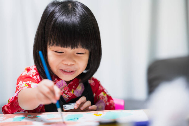 Szczęśliwy azjatyckie dziecko malowanie kolor wody na papierze sztuki. Słodki uśmiech dziewczyny z lekcją w szkole. Dzieciak w czarnym fartuchu. Pojęcie uczenia się zgodnie z preferencjami i uzdolnieniami. - Zdjęcie, obraz