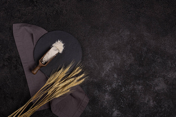 Белая пшеничная мука в деревянной ложке на темном структурном фоне на тарелке из черного камня. Уши пшеницы - Фото, изображение