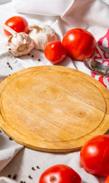 Une table avec différents tissus, sur laquelle repose une planche ronde, autour des tomates mûres rouges et de l'ail. Photo verticale. - Photo, image