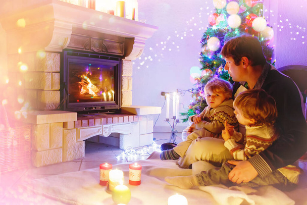 Père et deux petits garçons assis près de la cheminée, bougies et cheminée et regardant le feu. - Photo, image