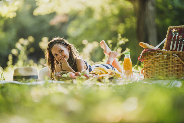 Urocza, uśmiechnięta dziewczynka leży na kocu na trawie i cieszy się dniem pikniku w parku. - Zdjęcie, obraz