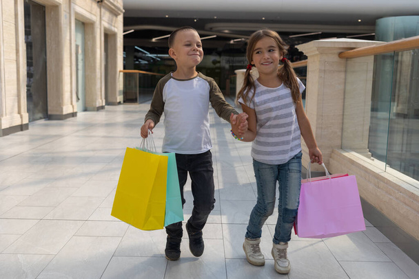 Portret grupy dwa cute adorable dzieci w wieku przedszkolnym zakupy. Kaukaski małej dziewczynki i chłopca w centrum handlowym. Dzieci trzymając torby na zakupy. - Zdjęcie, obraz