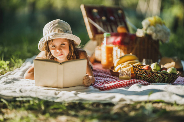 Ένα χαριτωμένο κοριτσάκι διαβάζει ένα βιβλίο ενώ απολαμβάνει την ημέρα πικνίκ στη φύση. - Φωτογραφία, εικόνα