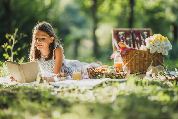 A cute uśmiechnięta dziewczynka czytając książkę podczas korzystania z piknik dzień w przyrodzie. - Zdjęcie, obraz