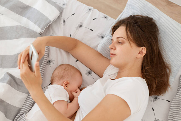Good looking tired sleepy woman wearing white casual style t shirt using cell phone for checking networks or typing messages while breast feeding her infant baby, lying bed. - Foto, afbeelding
